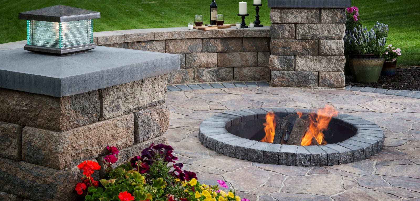 An outdoor stone patio with a fire pit, decorative plants, and a bottle of wine with glasses.