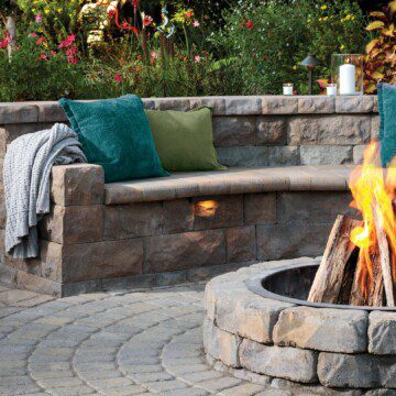 A stone bench curved around a fire pit; the bench has tidepool and seagrass colored throw pillows on it.