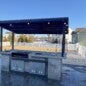 Outdoor kitchen installation with custom-built wood-burning grill, overhead cedar beams, stone fire pit, integrated bench seating, and happy client enjoying the finished space.