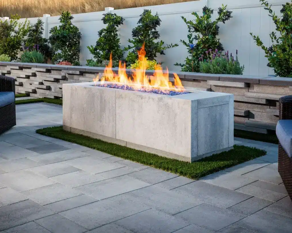 Outdoor patio with a rectangular concrete fire pit, decorative glass, and plants in the background.